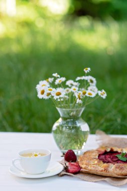 Taze pişmiş ev yapımı galet ya da açık çilekli turta, bir fincan bitki çayı ve bir buket papatya çiçekli vazo. Yumuşak seçici odak.