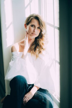 Kıvırcık ve dolgun dudaklı güzel seksi kız. Beyaz bluzlu, seksi kollu ve güneş ışığında pencerede kot pantolon giyen kız. Güzellik ve modaya uygun bir tarz. Yumuşak seçici odak.