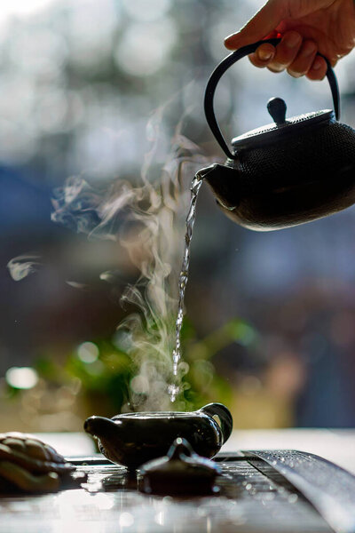 Tea ceremony. Hot tea in a teapot in hands, a glass and a cup on a background of a window. Soft focus.
