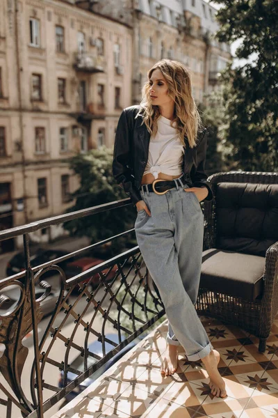 Şık, şık, göz makyajlı, kot pantolonlu, beyaz tişörtlü ve şehirde balkonda siyah deri ceketli bir kadın. Bahar sonbahar moda konsepti. Yumuşak seçici odak.