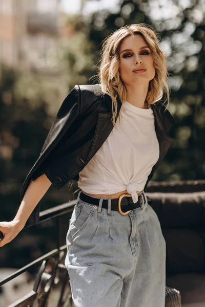 Şık, şık, göz makyajlı, kot pantolonlu, beyaz tişörtlü ve şehirde balkonda siyah deri ceketli bir kadın. Bahar sonbahar moda konsepti. Yumuşak seçici odak.