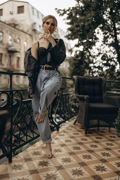 Şık, şık, göz makyajlı, kot pantolonlu, iç çamaşırlı ve şehirde balkonda siyah deri ceketli bir kadın. Bahar sonbahar moda konsepti. Yumuşak seçici odak.