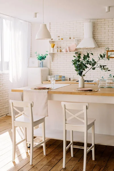 Modern interior of a white light kitchen in a Scandinavian style, a wooden table, natural linen textiles, a brick wall and kitchen utensils. Light loft. Soft selective focus.