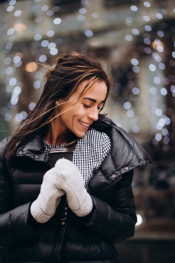 Eldivenli ve ceketli genç bir kadın, rüzgarlı bir kış gününde elinde bir fincan kahveyle Noel ışıklarının arka planına doğru gülümsüyor. Yumuşak seçici odak, sanat gürültüsü