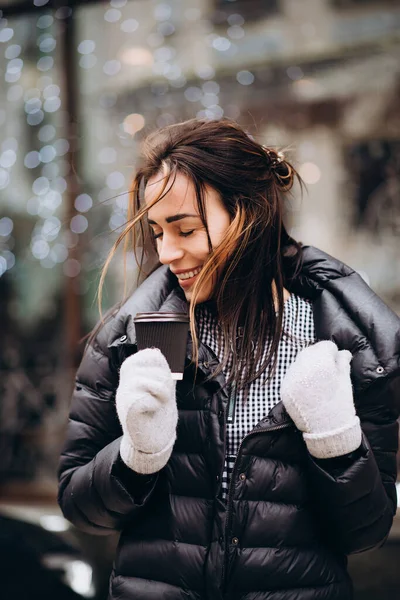ミトンの若い女性とダウンジャケットの笑顔と風の強い冬の日にクリスマスライトガーランドの背景に手にコーヒーのカップを保持しています ソフトフォーカス アートノイズ — ストック写真
