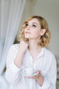 Aydınlık bir odada açık renk gömlekli güzel sarışın kız işe gitmeden önce bir fincan kahvenin tadını çıkarıyor. Banyodaki gizli elbiseli kız..