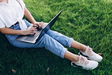 Bilgisayarlı kız öğrenci dışarıda çimlerin üzerinde oturuyor, internette sörf yapıyor ya da sınavlara hazırlanıyor. Teknoloji, eğitim ve uzaktan çalışma konsepti. Yumuşak seçici odak.