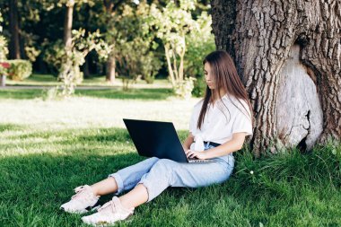 Diz üstü bilgisayarı olan bir kız öğrenci parktaki çimlerin üzerinde oturuyor, bir ağacın yanında, internette sörf yapıyor ya da sınavlara hazırlanıyor. Teknoloji, eğitim ve uzaktan çalışma konsepti. Yumuşak seçici odak.