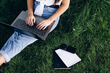 Bilgisayarlı kız öğrenci dışarıda çimlerin üzerinde oturuyor, internette sörf yapıyor ya da sınavlara hazırlanıyor. Teknoloji, eğitim ve uzaktan çalışma konsepti. Yumuşak seçici odak.