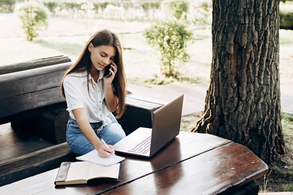 一个在户外带着笔记本电脑的漂亮的女学生坐在一张木制桌子旁 一边打电话一边聊天 教育和远程工作概念 软性选择性重点 — 图库照片