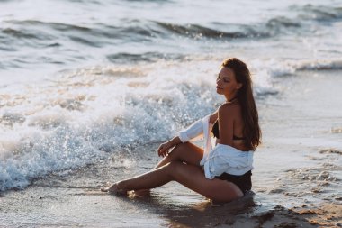 Siyah bikinili güzel bir kadın ve bronzlaşmış bir vücutta beyaz bir gömlek sahilde köpük dalgaları içinde oturuyor. Yumuşak seçici odak.