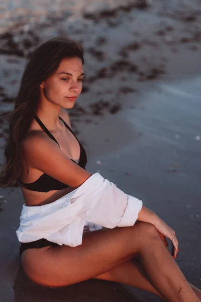 黒いビキニの若い美しい女性と日焼けした体の白いシャツは 湿った砂の上のビーチに座っています 柔らかい選択的フォーカス — ストック写真