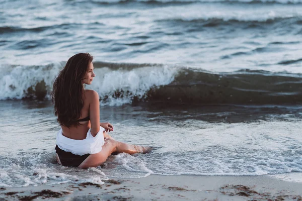A young beautiful woman in a black bikini and a white shirt on a tanned body sits on the beach in the waves. Soft selective focus.