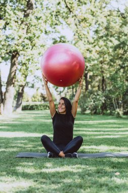 Genç, mutlu, bronz tenli esmer spor için büyük bir spor topuyla gelir, aktif siyah kıyafetler giyer, egzersiz yapar, spor yapar, parkta poz verir, dışarı çıkar. Yumuşak seçici odak.
