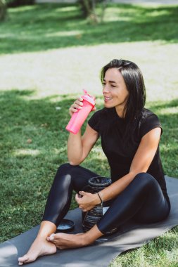 Bronz tenli genç bayan sporcu, açık hava eğitiminden sonra protein içeceği içiyor. Spor beslenmesi. Yumuşak seçici odak.