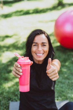 Bronz tenli genç bayan sporcu, açık hava eğitiminden sonra protein içeceği içiyor. Spor beslenmesi. Yumuşak seçici odak.