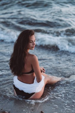 Siyah bikinili güzel bir kadın ve bronzlaşmış bir vücutta beyaz bir gömlek dalgaların arasında kumsalda oturuyor. Yumuşak seçici odak, sanat burnu.