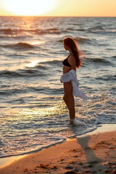 Plajda bronzlaşmış bir vücutta siyah bikinili ve beyaz gömlekli güzel bir kadın gün batımına hayran. Yumuşak seçici odak, sanat burnu.
