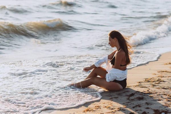 A young beautiful woman in a black bikini and a white shirt on a tanned body sits on the beach in waves of foam. Soft selective focus, art nose.
