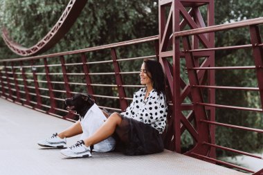 Pofuduk etekli ve benekli bluzlu güzel bir kadın köprüde Staffordshire Bull Terrier köpeğiyle oturuyor. Yumuşak seçici odaklanma. Sanatsal gürültü.