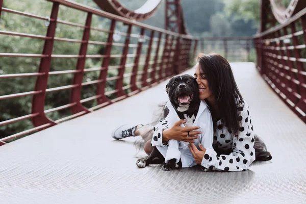 A beautiful young woman in a fluffy skirt and a polka-dot blouse sits on the bridge and hugs her Staffordshire Bull Terrier dog. Soft selective focus. Artistic noise.