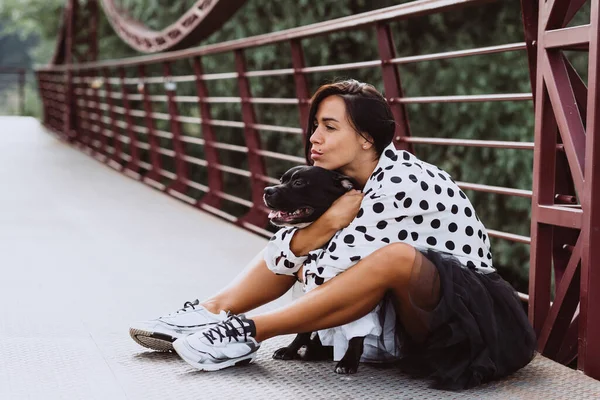 A beautiful young woman in a fluffy skirt and a polka-dot blouse sits on the bridge and hugs her Staffordshire Bull Terrier dog. Soft selective focus. Artistic noise.