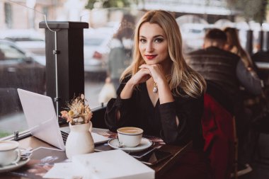 Dizüstü bilgisayarı olan genç ve güzel bir iş kadını sonbaharda öğle yemeği arasında bir kafede bir fincan kahvenin keyfini çıkarıyor. Yumuşak seçici odak, sanatsal ses.