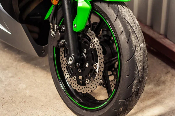 Front wheel with brake discs, caliper of green sports motorcycle close-up on concrete. Rubber with black alloy disc, brake system of road bike in garage on sale. Moto tire and fork. Front side view.