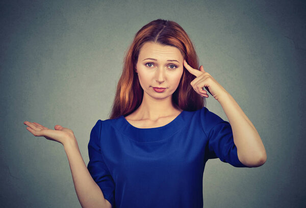 Красивая рыжая девушка скручивает палец в висок и смотрит в разногласиях на камеру на сером фоне
