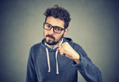 Sakallı genç adam yumruk up ciddi seyir ve tehdit ile gri arka plan üzerinde tutarak gözlük. 