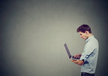 Yan görünüm rahat genç adam tutarak ve modern dizüstü ayakta gri arka plan üzerinde kullanma