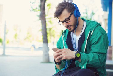 Trendy delikanlı Caddesi üzerinde otururken akıllı telefon ve kulaklık ile müzik dinlemek