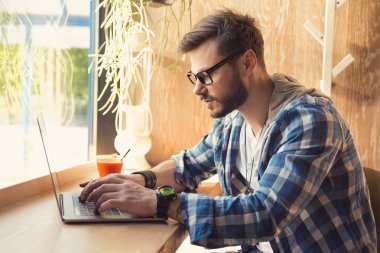 Genç hipster adam içki içiyordum ve güneş ışığı altında dizüstü bilgisayar kullanarak kafeteryasında freelancing gözlük