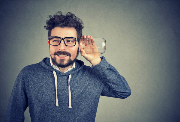 Молодой любопытный человек держит стеклянную банку возле уха и слушает сплетни с волнением на сером фоне
