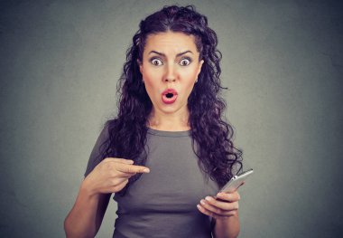 Genç kadın kamera akıllı telefon gri arka plan üzerinde izole Web sayfasında şok haber okuduktan sonra işaret dehşet ifade ile seyir. 