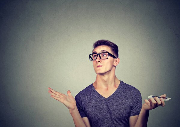 Young casual man in glasses holding smartphone and looking up displeased with conversation on gray background