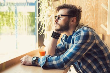 Hipster genç adam tek başına kafeterya uzakta penceresinde üzüntü bakarak oturmak bardaklarda yan görünüm