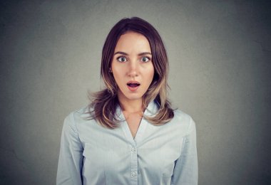 Stunned woman in blue shirt standing with mouth opened and looking at camera on gray background clipart