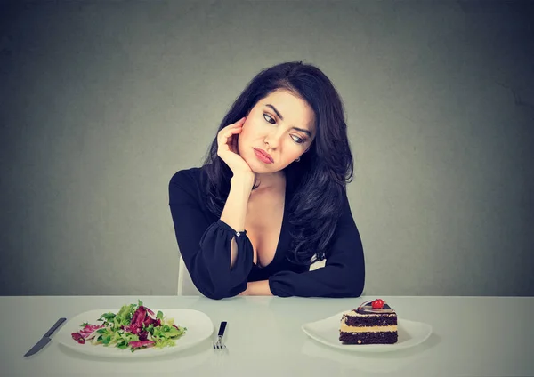 サラダ ケーキ デザートのための渇望と灰色の背景にダイエットのジレンマを持つテーブルに座っていた若い女性 — ストック写真