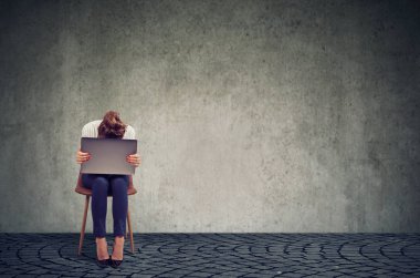 Diz üstünde laptop ile sandalyede oturan ve kriz gri duvar arka plan karşı umutsuz seyir anonim kadın