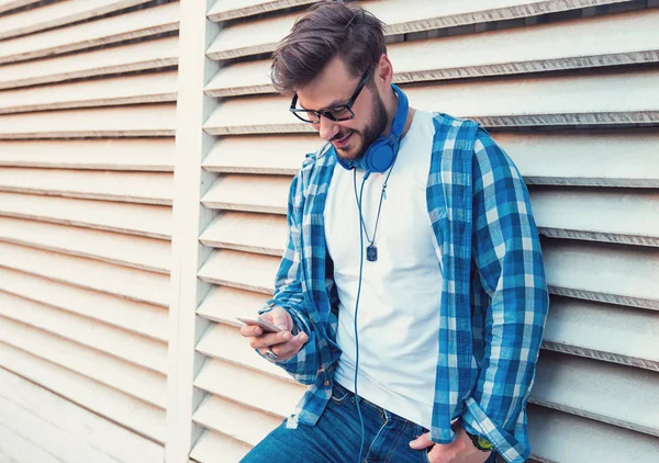 青いシャツと路上の壁にもたれながら笑みを浮かべてスマート フォンをサーフィンのヘッドフォンでトレンディーな流行に敏感な男 — ストック写真