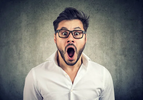 Beyaz gömlek giymiş rahat genç adam görünüyor dinleme cihazı gözlerle, gri arka plan üzerine açık ağızlı şok hayran. 