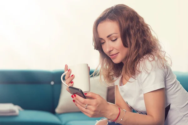 Jovem Mulher Sentada Sofá Usando Smartphone Enquanto Toma Café Luz — Fotografia de Stock