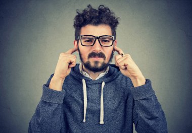 Kameraya huysuz seyir ve gürültü gürültü ve çakışma durumu gri arka plan üzerinde kaçınarak kulaklarından kapsayan rahatsız genç hipster adam 