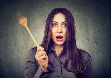 Young housewife holding wooden spoon and looking with great surprise and shock at camera on gray background clipart