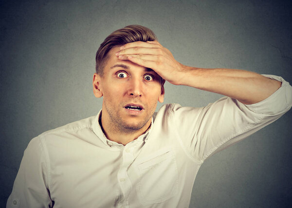 Взрослый мужчина в белой рубашке держит руку на лбу и выглядит супер пораженным камерой на сером фоне
