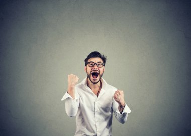 Super excited young man in glasses holding fists in celebration of success on gray background clipart