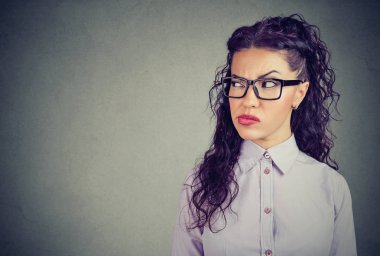 Young curly brunette in glasses looking away in jealousy making revenge plan on gray background clipart