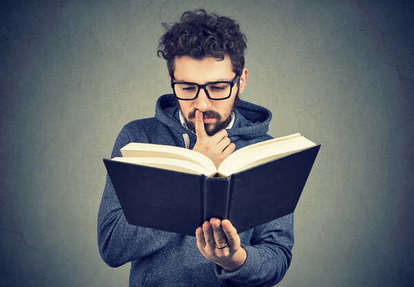 Молодой человек в очках выглядит смущенным, пытаясь прочитать умную книгу на сером фоне
