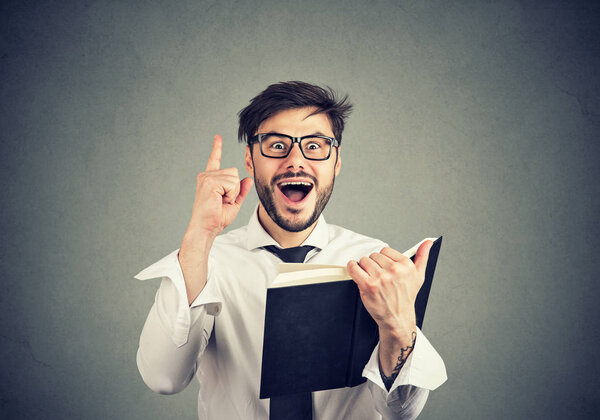 Молодой умный бизнесмен в очках, держа палец вверх взволнован отличной идеей, держа книгу на сером фоне
